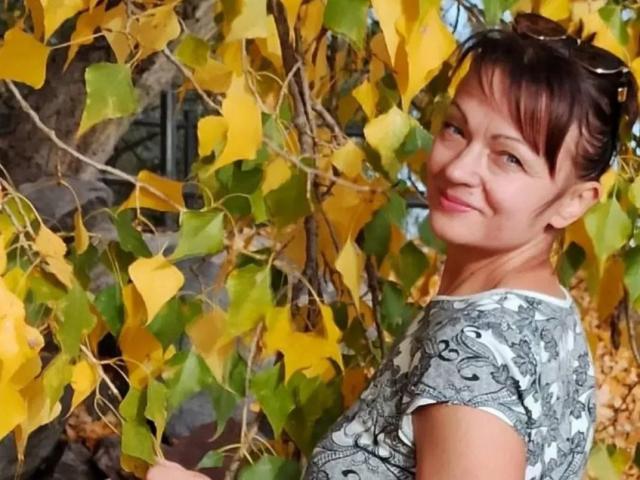 AuroraRosco with short brown hair, wearing a gray patterned top, smiling, standing in front of yellow and green autumn leaves, sunglasses on her head.