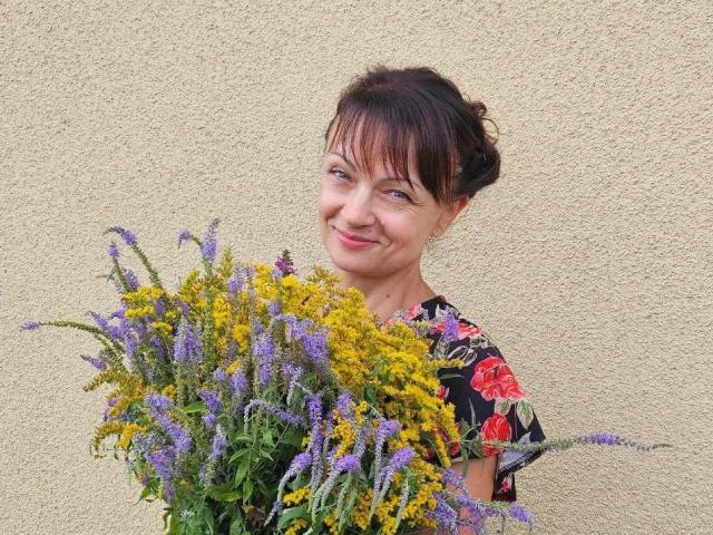 AuroraRosco with a smile, holding a bouquet of yellow and purple flowers, wearing a floral dress, standing against a textured beige wall. Her tits are partially visible.
