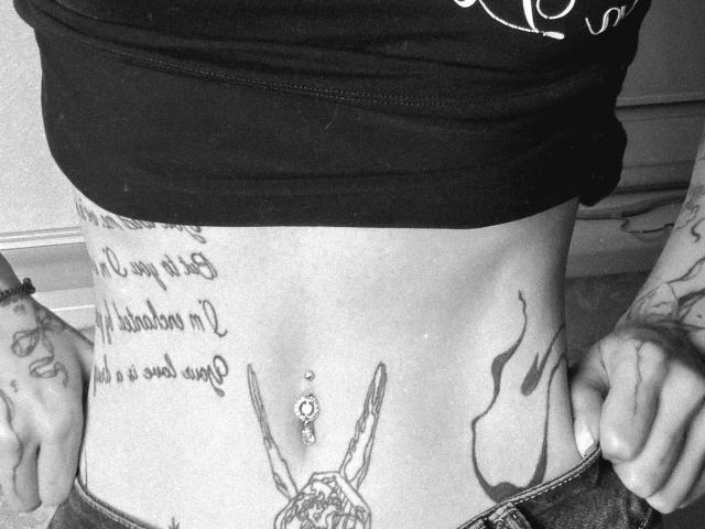 SensualMila's flat, tattooed, black-shirted midriff, showing a navel piercing and wing tattoos, with hands pulling down jeans, revealing a belly button and lower torso tattoos.