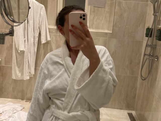 Dionisia in a white fluffy bathrobe, taking a selfie with her phone, covering her face. She has red nails and is in a tiled bathroom.