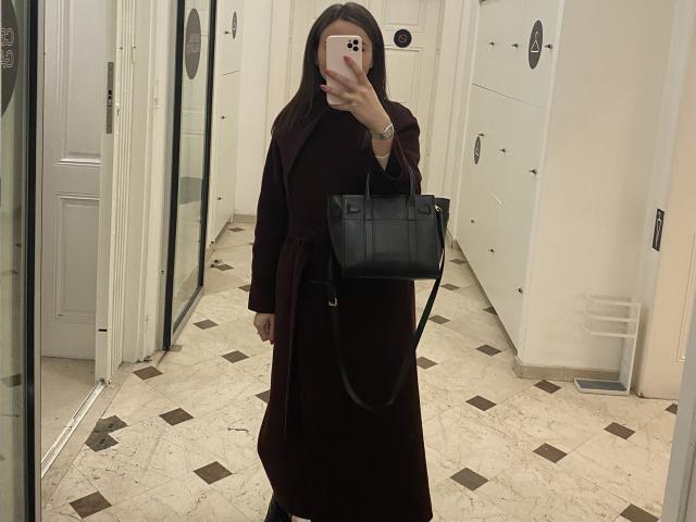 Dionisia with long dark hair, wearing a brown long dress and black handbag, holding a phone, covering her face, standing in a white-walled, marble-tiled hallway.
