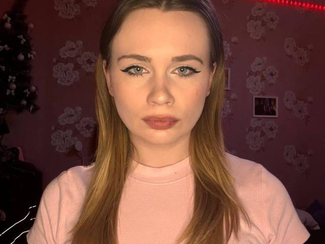 CheriDahmen hat langes, blondes Haar, trägt ein pinkes Hemd, hat schwarzen Eyeliner, pinken Lippenstift und einen ernsten Gesichtsausdruck. Im Hintergrund befindet sich eine rote LED-Leiste..