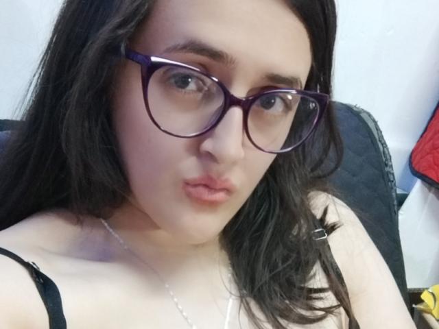 AndreaHotLove69 with dark brown hair, purple glasses, and pouty lips, wearing a black strap top, showing pale skin and a slight cleavage.