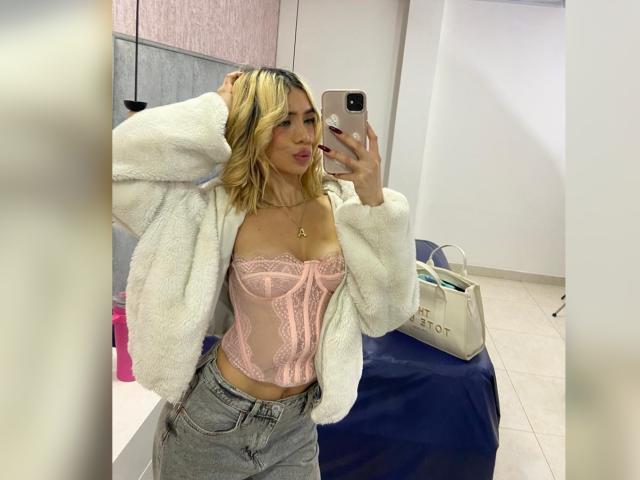 ChristieGarci with blonde hair, pink lace-up corset, white fuzzy jacket, and gray jeans, taking a mirror selfie with her phone, showing her tits and small ass.