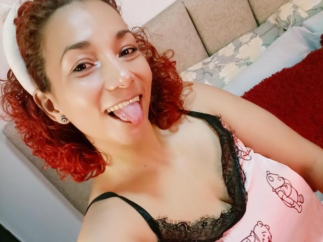 AmberDemons with curly red hair, tongue out, wearing a pink nightie with black lace and cartoon prints, has small breasts, and a white headband in a bedroom.