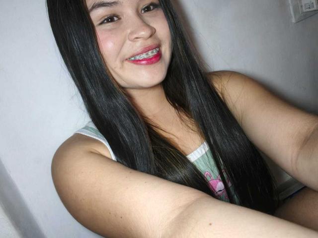 NaughtyMissyy with long black hair, smiling with pink lipstick, showing metal braces, wearing a green tank top, taking a selfie with her right arm.