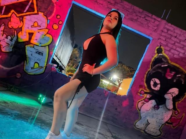 NaughtyMissyy in a black, halter-neck, cut-out dress standing with her ass facing the camera, legs in a pool, neon lights, graffiti wall, night, and blue water.