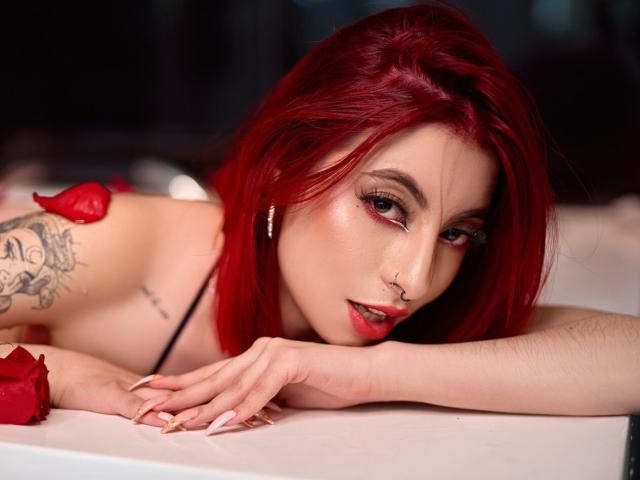 SophieMaiden with red hair, nose ring, and tattoos, lying on white surface, red rose petals, looking seductively at camera, showing off her tits and sexy, pale skin.