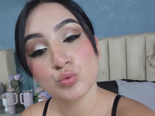 HiilaryDolce with pouty lips, wearing black eyeliner, shimmering eyeshadow, pink blush, and black top, her dark hair tied back, blue wall in background.