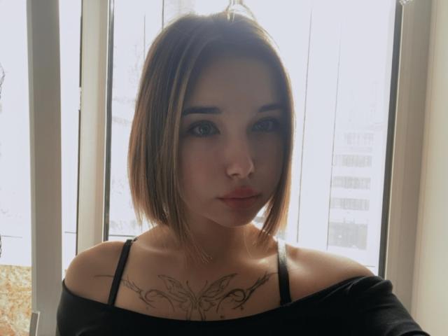 LavetaGurule has short brown hair, black off-shoulder top, and a butterfly tattoo on her chest. She's looking at the camera with serious eyes, standing in front of a window.