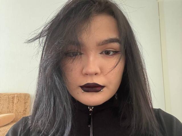 ElenaDecuir, avec du rouge à lèvres foncé, de l'eye-liner noir et des cheveux noirs, a un piercing au nez et un piercing à la lèvre inférieure, elle porte une veste noire zippée et regarde directement l'appareil photo..