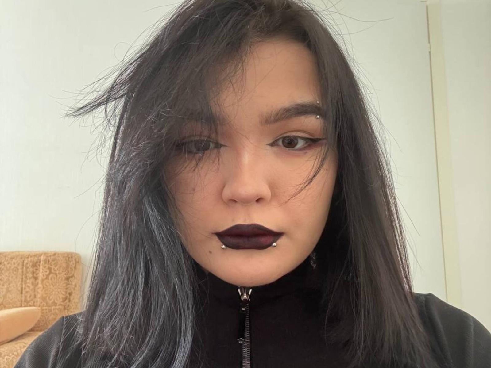 Elena Decuir, mit dunklem Lippenstift, schwarzem Eyeliner und schwarzen Haaren, hat ein Nasenpiercing und ein Piercing im Unterlippenbereich, trägt eine schwarze Jacke mit Reißverschluss und blickt direkt in die Kamera..