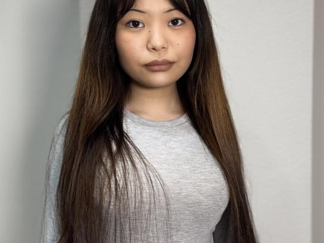 Joetta Bealer mit langen, braunen Haaren, einem grauen Hemd und einem hübschen Gesicht, die kleine Brüste zeigt und einen ruhigen Ausdruck hat. Eine asiatische Frau mit glatten Haaren, neutralem Make-up und einem leichten Lächeln..