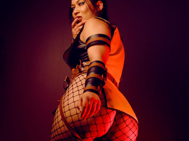 KendallBrzz with a sexy, curvy ass, wearing fishnet stockings, black leather straps, and a tan coat, biting her finger, looking over her shoulder, in a red-lit photo.