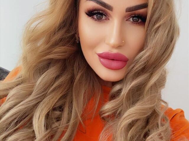 NicolSNaya has blonde, wavy hair, thick eyebrows, big eyelashes, and pink lipstick. She's wearing an orange top, and her nose is pierced on the right side.