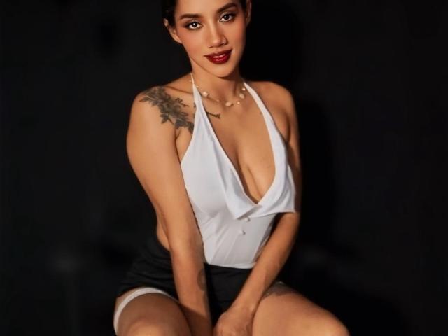 LunaGomezz with big boobs, wearing a white halter top, black shorts, and a necklace, sitting with hands on knees, black background, tattoo on right shoulder, red lipstick.