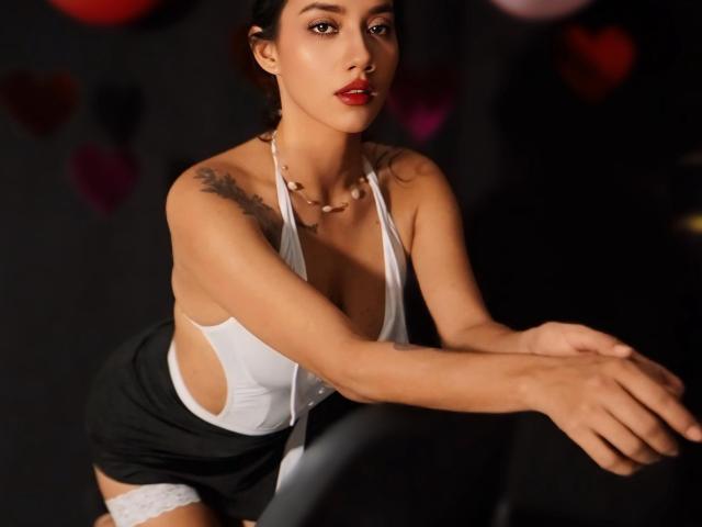 LunaGomezz, a woman with olive skin and dark hair, leans forward with arms extended. She wears a white, deep-cut halter top, revealing cleavage and side tattoos. A black skirt and white lace thigh-highs complement her outfit. Red lipstick contrasts with her serious expression. She has a necklace with small beads. The background is dark with blurred heart shapes. The image focuses on her upper body and face, highlighting her tattoos and outfit.