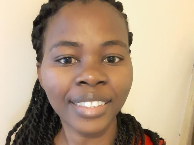 Close-up of Chalaah with dark brown skin, black twisted braids, and a subtle smile. Chalaah has medium-sized, symmetrical brown eyes and thin, arched eyebrows. The white teeth are slightly spaced. Chalaah wears a red garment with a black pattern partially visible. The lighting is even, highlighting the smooth texture of Chalaah's skin. The background is plain and off-white. Chalaah's expression is neutral, with a slight upward curve at the corners of the mouth.