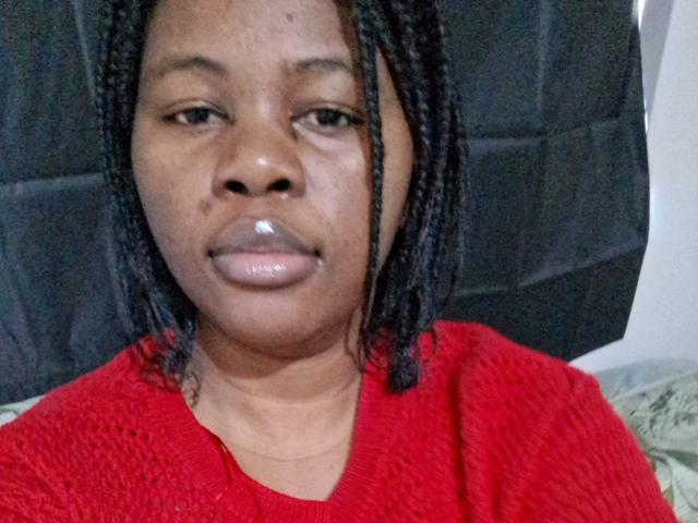 Chalaah has shiny lips, dark braided hair, and is wearing a red sweater. Her face is centered, showing a neutral expression. She's a dark-skinned woman with a chubby build.