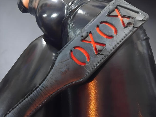 Los pantalones de látex negro brillante de Carolaine Foster, con un "OXO" rojo grabado en un cinturón de cuero negro, que muestran sus curvas y su piel lisa y reflectante..