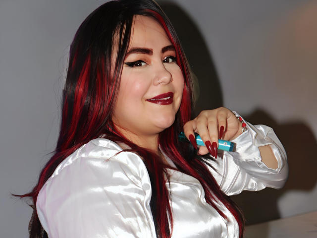 Carolaine Foster, con capelli rossi e neri, una camicia di seta bianca, rossetto scuro e unghie lunghe rosse, tiene in mano una penna blu e sorride leggermente. Ha un viso carino e formoso..