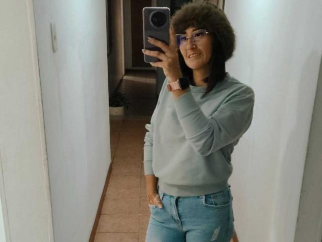 A photograph of SharissWillian standing in a hallway. She has medium-length brown hair and is wearing glasses. She's dressed in a light gray sweater and blue jeans with her right hand in her pocket. She holds a smartphone in her left hand, taking a mirror selfie. Her skin tone is medium, and she is smiling slightly. She wears a red smartwatch on her left wrist. The hallway has white walls and pink tiled floor.