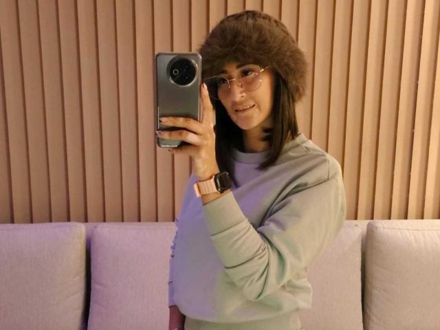 A selfie of SharissWillian with a smartphone. She wears a brown fur hat, glasses, and a light grey sweater. Her brown hair is straight, and she smiles slightly. She holds the phone in her right hand, showing a gold watch on her left wrist. The background has vertical wooden slats and a beige couch. The composition is casual and modern.