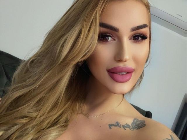 NicolSNaya has blonde hair, dark eyebrows, pink lipstick, and a tattoo on her right shoulder. She's wearing a necklace and has long, wavy hair covering her left side.