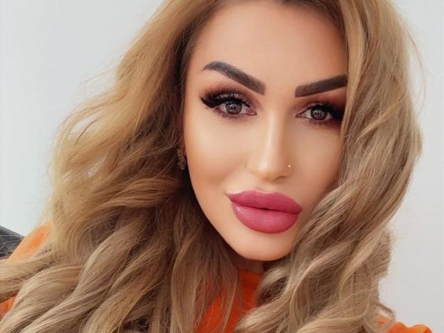 NicolSNaya has wavy blonde hair, thick eyebrows, long eyelashes, and bright pink lipstick. She's wearing an orange top with a small nose ring on her right side.
