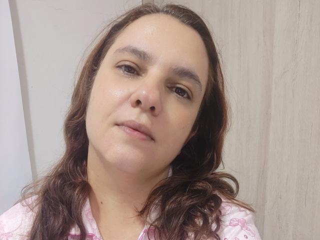AmeliaWilson has wavy brown hair, light skin, and subtle makeup with brown eyes. She wears a pink and white patterned shirt, looking slightly tired with a neutral expression.