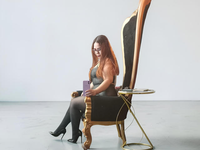 CarolaineFoster with orange hair, glasses, black boots, and gray top, sitting on gold and black throne, holding purple tablet, right leg crossed, small gold table to the right.