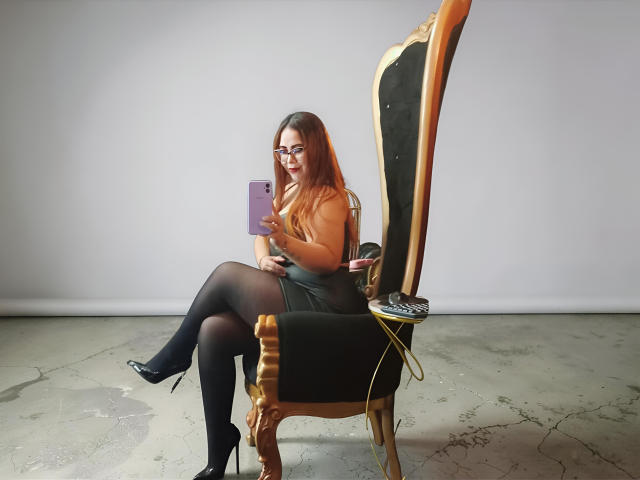 CarolaineFoster with long red hair, glasses, black skirt, black stockings, high heels, sitting on ornate chair, holding purple phone, cracking floor, white wall.