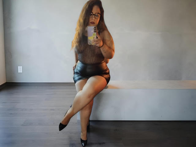 CarolaineFoster with wavy brown hair, glasses, sheer black top, black leather shorts, black heels, sitting on bench, taking mirror selfie, showing her ass and legs.