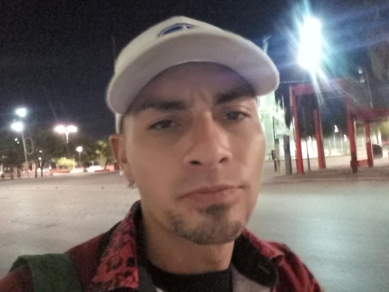 Das Gesicht von GeorgeDoo, mit einer weißen Mütze, einer roten Jacke mit schwarzem Besatz und einem grünen Rucksack, mit einem hellbraunen Bart, steht in einem schlecht beleuchteten Außenbereich bei Nacht..
