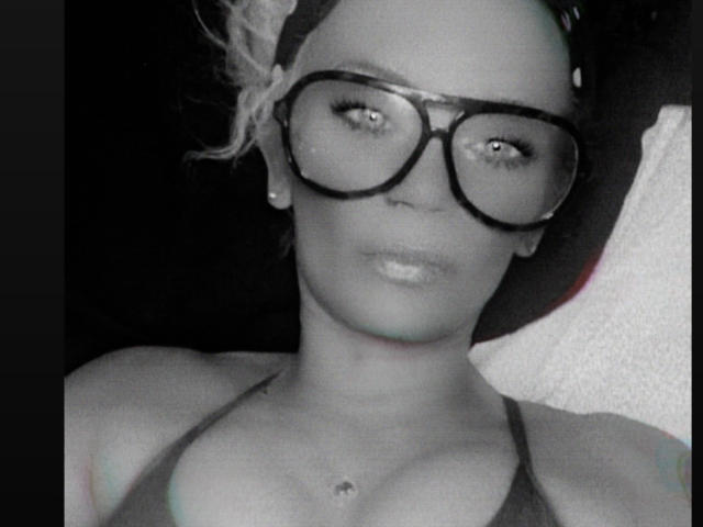 Selfie en blanco y negro de SnowBelly con gafas grandes, oscuras y redondas, una diadema y un top de tirantes, mostrando sus pechos y un collar pequeño sobre un fondo oscuro..