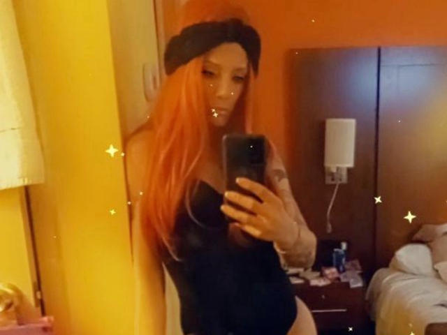 SnowBelly com cabelos longos e laranja, boina preta, top preto sem alças, segurando um telefone, selfie no espelho, quarto amarelo, cama visível, abajur pequeno e mesa de cabeceira, mostrando seus seios e nádegas..