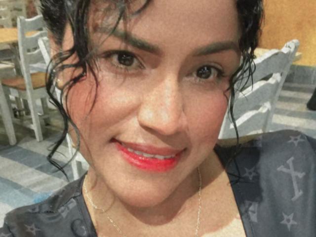 IvannaDuran has curly black hair, green eyebrows, and red lipstick, smiling with white teeth. She's wearing a grey shirt with star patterns and a silver necklace.