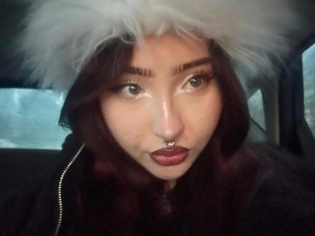 AriaVerlangen-hot with dark maroon hair, white fluffy hat, silver nose ring, and dark lipstick, looking at the camera with green eyes in a car.