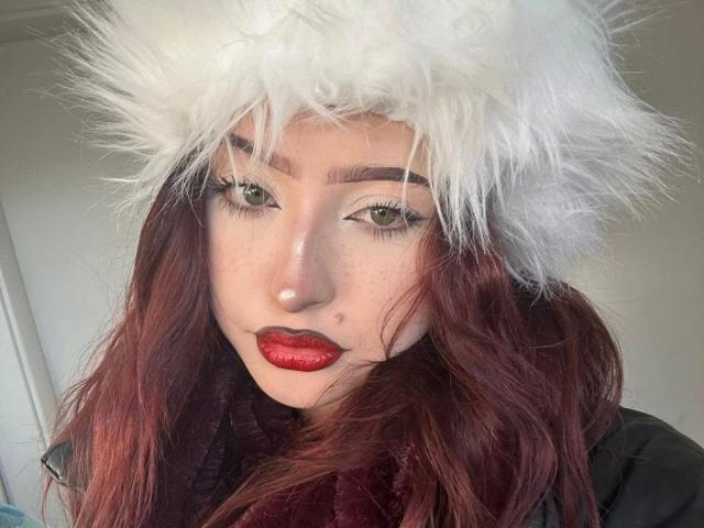 AriaVerlangen-hot has fair skin with freckles and wears red lipstick. She has green eyes with long, dark eyelashes. Her wavy, red-brown hair cascades down. She wears a fluffy white hat and a dark scarf. Her eyebrows are thick and arched. The image focuses on her face, showing a confident, direct gaze. The texture of the hat contrasts with her smooth skin. She has a small mole on her right cheek. The lighting is natural, highlighting her features.