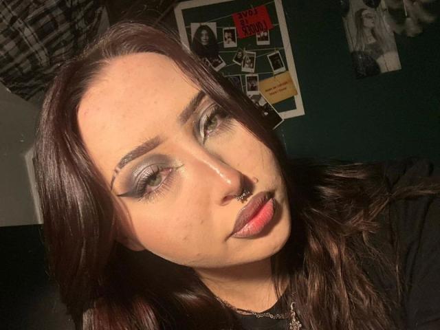 AriaVerlangen-hot has long, dark brown hair, green eyes with heavy gray eyeshadow, and a small silver nose ring. Her dark pink lipstick is slightly glossy. She has a fair complexion with light freckles and a subtle blemish on her cheek. She wears a black top. The background shows a dark green wall with a poster featuring black-and-white photos. The lighting is warm, casting shadows on her face. Her expression is neutral, looking slightly to the right. The overall style is casual with a focus on makeup.