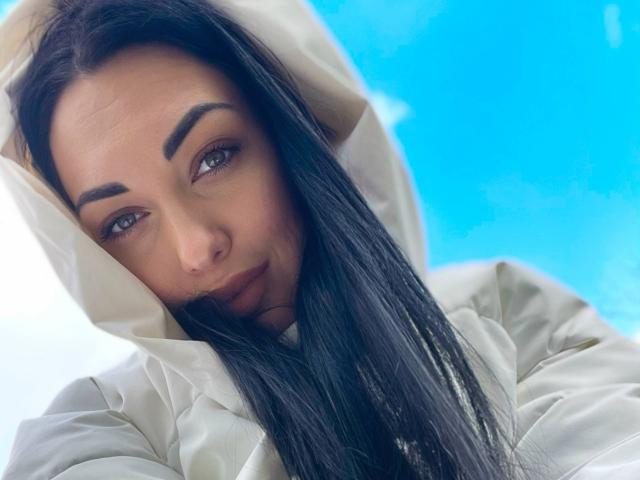 Xioni with long black hair, brown eyebrows, and dark lipstick, wearing a white hood, looking down with a slight smile. Bright blue background. Soft, feminine features.