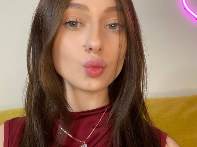Close-up of TherealMissa with long brown hair, slightly parted lips, and a small nose stud. She has light skin, blue eyes with long lashes, and a subtle pink lip color. She wears a sleeveless maroon top and a thin gold necklace with a small pendant. Background includes a yellow surface and a faint purple neon sign. Her expression is neutral, almost pouting. The image is sharp with natural lighting.