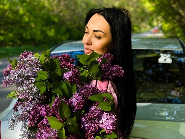 Xioni with long black hair holding a large bouquet of purple and white lilacs in front of a silver car. Her eyes are closed, and she's wearing a pink shirt.