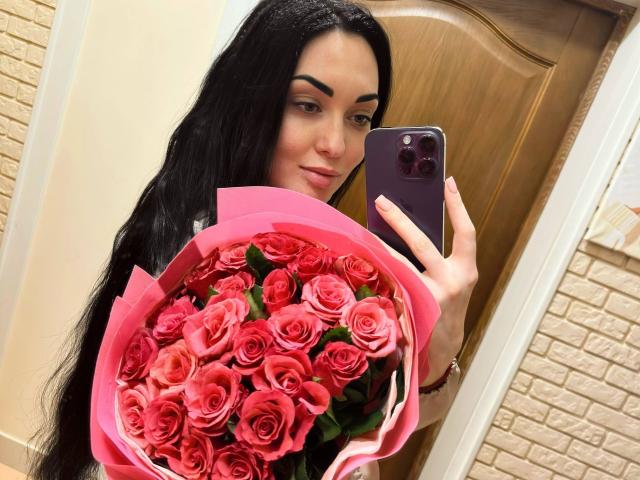 Xioni with long black hair, holding a pink bouquet of red roses in front of a mirror, taking a selfie with a purple phone, smiling slightly.