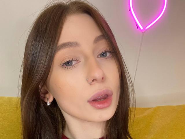 TherealMissa has pale skin, brown hair, blue eyes, a nose ring, and a small hoop earring. She's wearing pink lipstick and has a shiny, pouty expression. Neon pink heart.