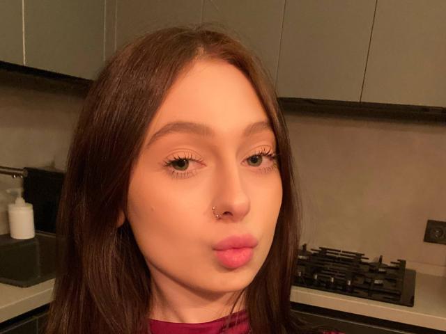 TherealMissa has long brown hair, green eyes, a nose piercing, and pink lips, making a pouty face in a kitchen with grey cabinets and a gas stove.
