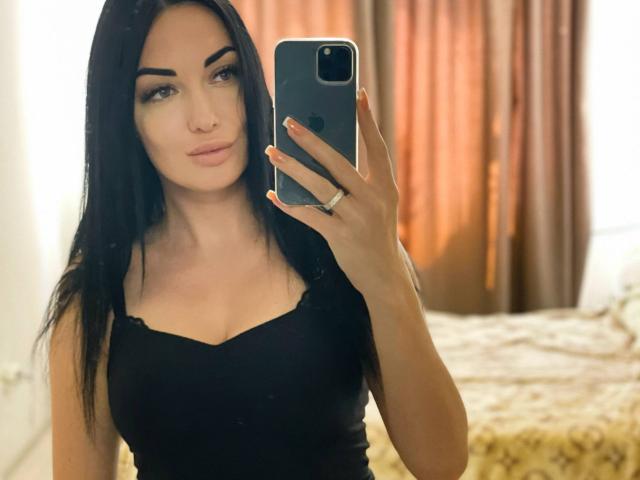 Xioni, avec de longs cheveux noirs, portant un haut noir, tenant un téléphone pour un selfie devant un miroir, montrant ses seins, et regardant l'appareil photo avec un léger sourire..