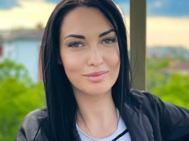 Xioni has black hair, green eyes, and a subtle smile. She's wearing a black jacket over a white shirt. Her eyebrows are dark and well-defined.