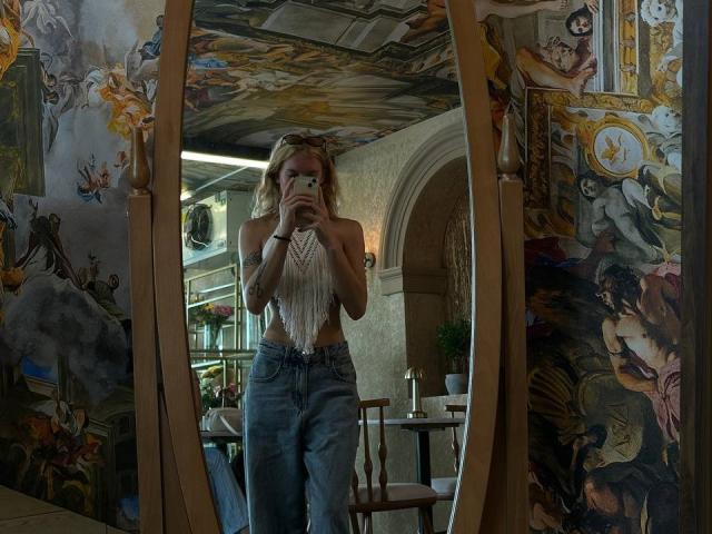YaekoBordges, reflected in a wooden mirror, takes a selfie. She has blonde hair, a fringe, and wears a fringed white top with high-waisted blue jeans. The background features ornate, colorful paintings with religious and mythological scenes. The mirror frame is wooden, and the room has a mix of natural and artificial light. The mirror is centered in the image, capturing YaekoBordges from the waist up.