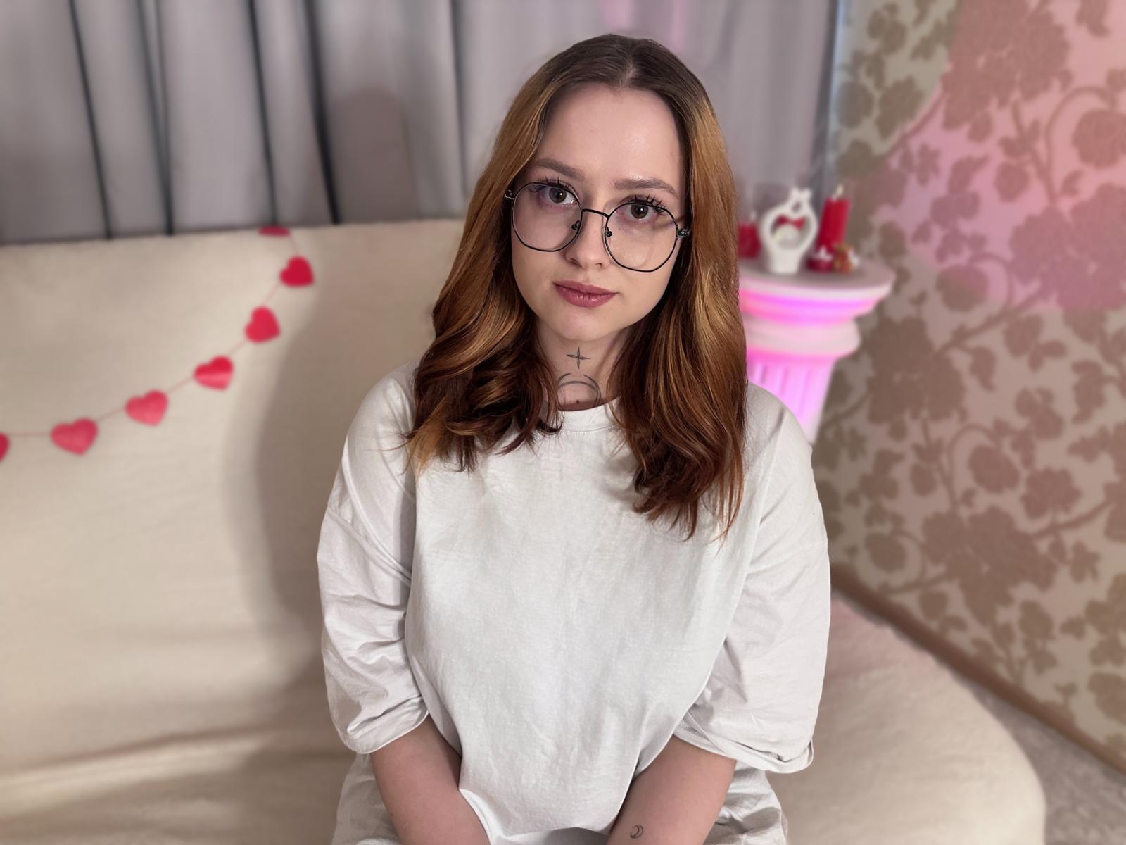 PolySoft, une jeune fille mignonne avec des lunettes et des cheveux bruns ondulés, portant une chemise blanche, est assise sur un canapé beige avec des décorations en forme de cœurs roses et une petite table rose..